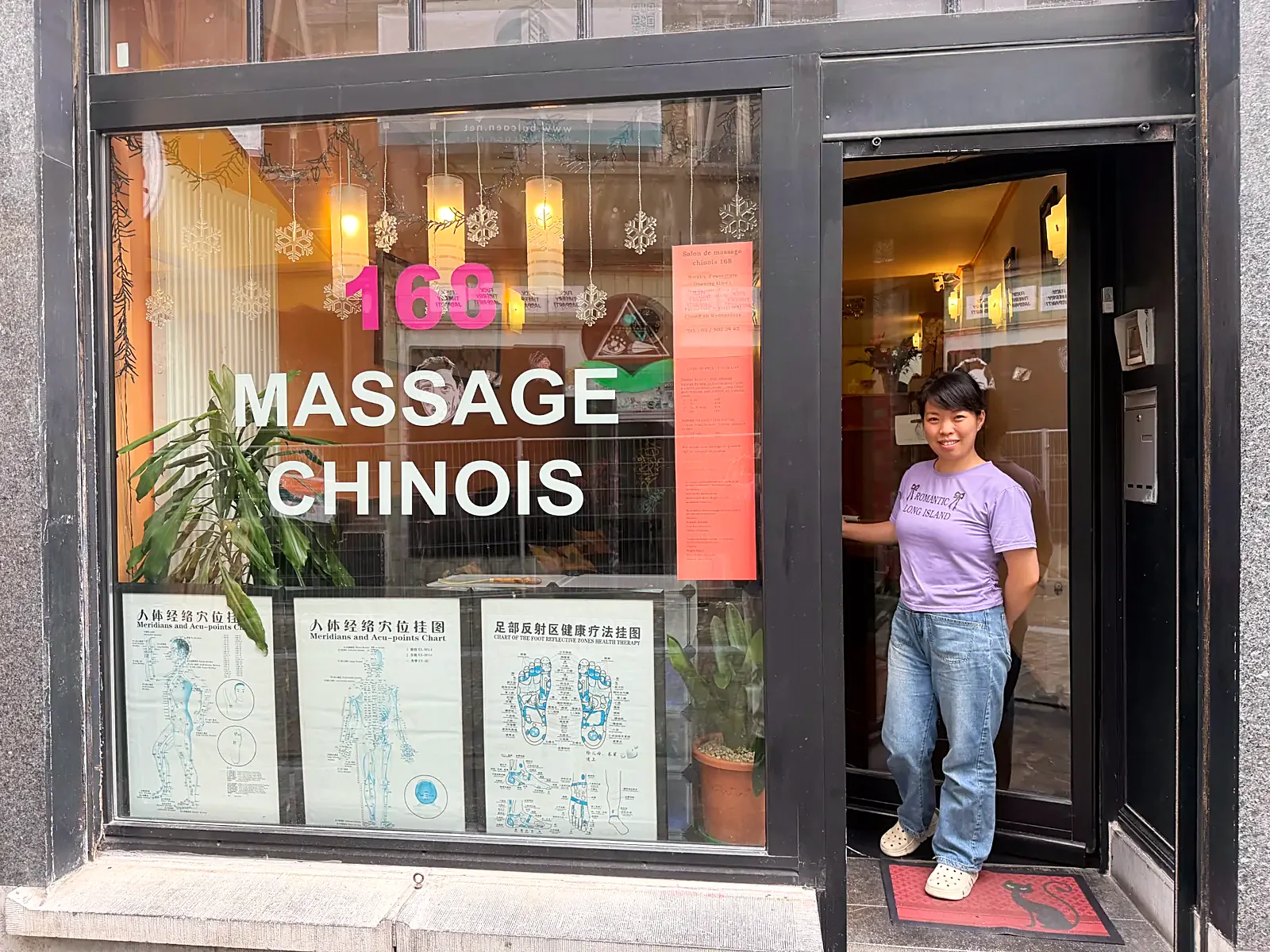 HaiHong vous accueille à l'entrée du centre, Rue des Chapeliers 32 à Bruxelles HaiHong, praticienne de massage chinois, souriante sur le seuil du centre Massage Chinois 168, Rue des Chapeliers 32 à Bruxelles, devant la vitrine et les planches d'acupression